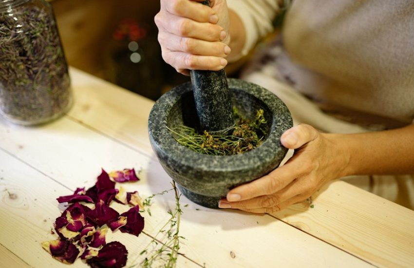 A person grinding herbal ingredients in a stone mortar and pestle with dried petals nearby.